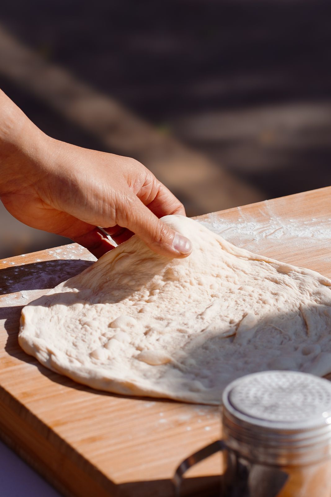 A hand stretches dough on a floured board in bright natural light.
