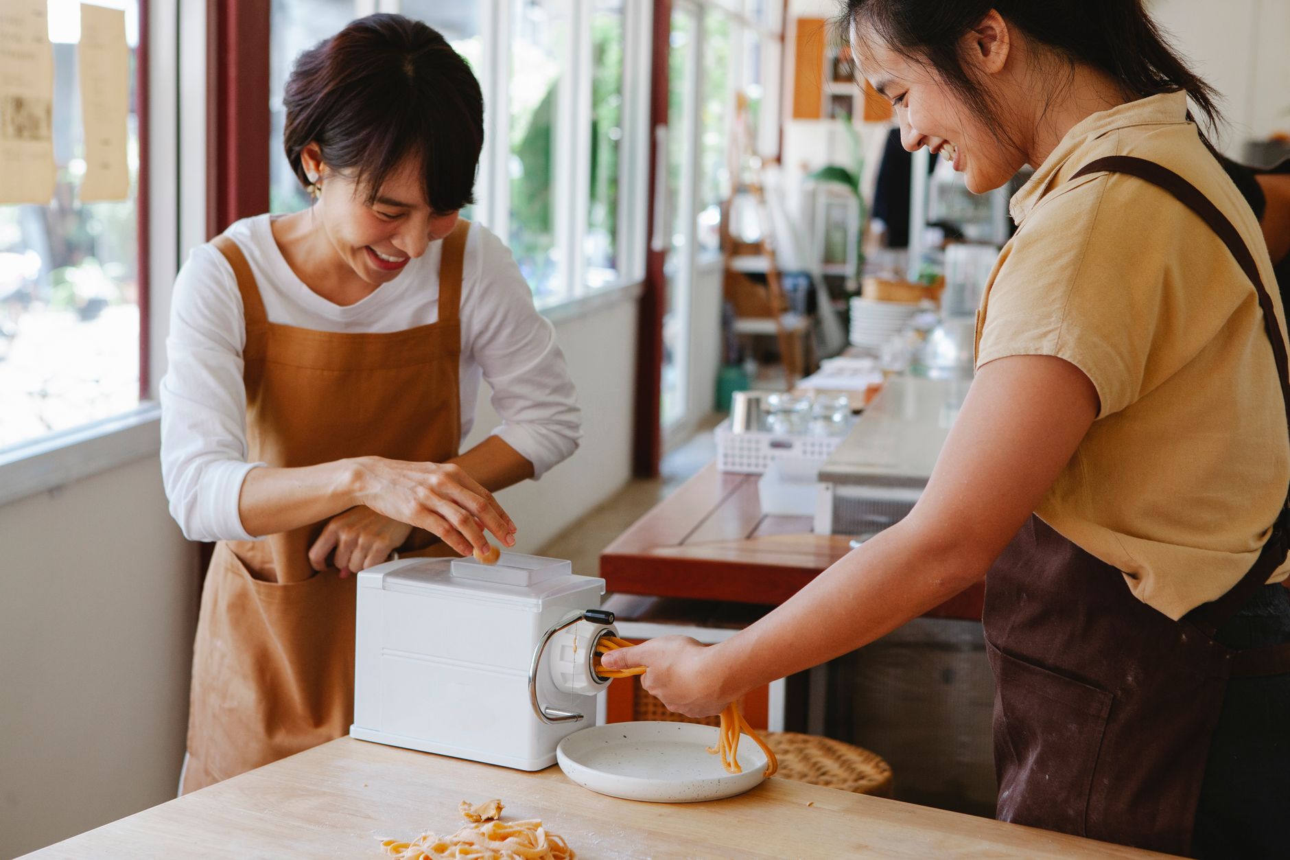 Two women guide fresh noodles through a pasta machine in a sunlit kitchen.